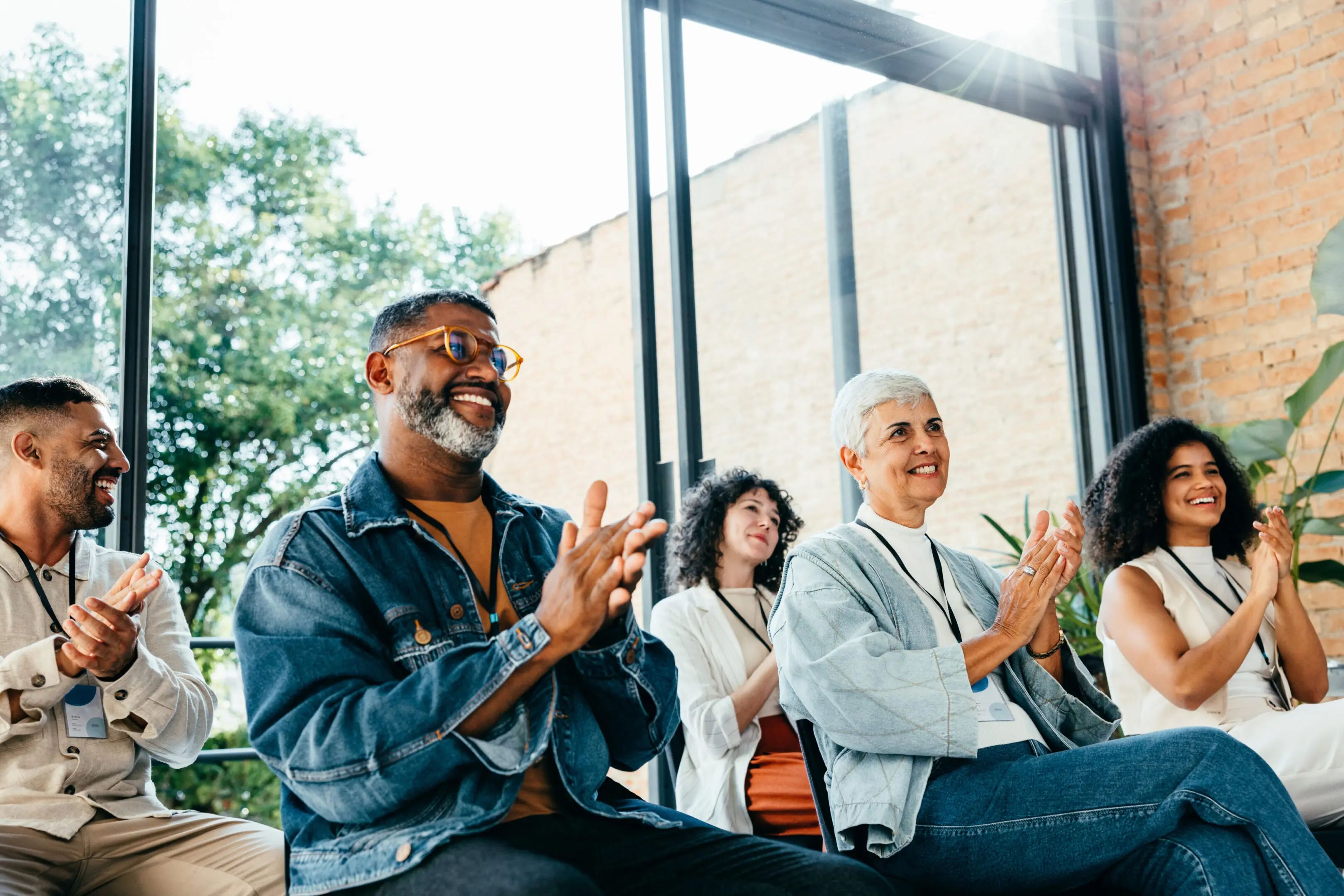 Diverse group of people attending a business meeting, happily clapping and smiling Diverse group of people attending a business meeting, happily clapping and smiling. They are seated in a bright room with large windows and natural light.